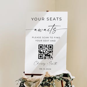 以下が含まれることがあります： 木製のイーゼルに立てられた白いウェディングサイン。テキストは「YOUR SEATS awaits」と書かれています。QRコードと「Chrissy + Smith」の名前と日付が含まれています。サインは緑と布で飾られています。