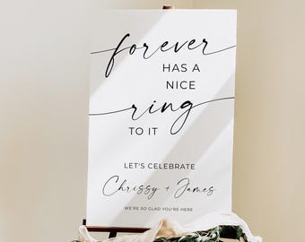 May include: A white sign on a wooden easel with the text "forever has a nice ring to it." Below, it reads "Let's celebrate Chrissy + James." The sign is decorated with greenery and fabric.