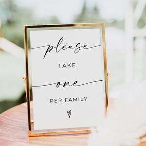 May include: A gold-framed sign with the words "please take one per family" in black script. The sign is on a wooden surface, with a blurred background. A decorative feather is in the foreground.