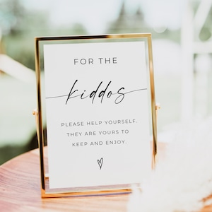 May include: A gold framed sign with the text "FOR THE kiddos" in a script font. Below, it reads "PLEASE HELP YOURSELF. THEY ARE YOURS TO KEEP AND ENJOY." A heart symbol is at the bottom. The sign is on a wooden surface.