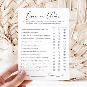 May include: A white card with the text "Over or Under" in elegant script, designed for a wedding game. The card features a series of questions with spaces for "under," "over," and "correct" answers. The card is held by a hand.