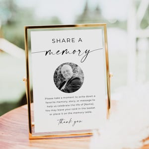 May include: A gold-framed sign with the words "SHARE A memory" in elegant script. Below, a black and white photo of a man is centered. The sign encourages guests to write a memory and includes a "Thank you" message.