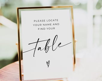 May include: A gold-framed table sign with the text "PLEASE LOCATE YOUR NAME AND FIND YOUR table" in black script. A small heart is below the word "table". The sign is on a wooden surface.