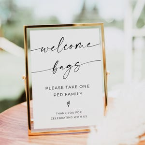 May include: A framed sign with gold trim and a white background. The sign reads "welcome bags" in elegant script, followed by "PLEASE TAKE ONE PER FAMILY" and "THANK YOU FOR CELEBRATING WITH US."
