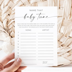 May include: A white baby shower game card with the text "Name That Baby Tune" in a handwritten font. The card has lines for song and artist names. The card is held in front of a woven beige background.