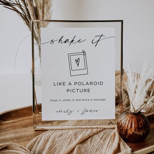 Puede incluir: Un cartel con marco dorado con el texto "shake it" en cursiva, un dibujo de una cámara Polaroid con un corazón dentro, y el texto "LIKE A POLAROID PICTURE" en mayúsculas. Debajo de la imagen está el texto "Snap it, shake it and leave a message." y los nombres "emily + Jones" en cursiva.