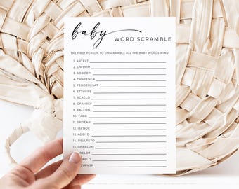 May include: A white baby shower word scramble game card with the word "baby" in cursive. The card has a list of scrambled words to unscramble. The card is held in front of a woven beige background.