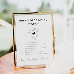 May include: A framed sign with gold trim displays instructions for a onesie decorating station. The sign reads "ONESIE DECORATING STATION" and includes a graphic of a onesie with a heart. Instructions are provided for applying vinyl designs.