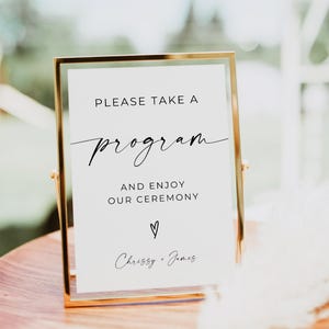 May include: A gold-framed sign with the text "PLEASE TAKE A program AND ENJOY OUR CEREMONY" in black script. The sign also includes a heart symbol and the names "Chrissy + James". The sign is on a wooden surface.