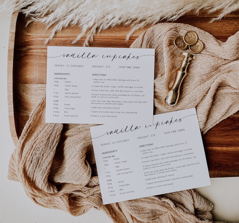 K&ouml;nnte beinhalten: Zwei Rezeptkarten f&uuml;r Vanille-Cupcakes werden auf einem Holztablett mit einem beigen Tuch pr&auml;sentiert. Die Karten enthalten handschriftlichen Text mit Zutaten und Anweisungen. Ein goldfarbener Wachssiegelstempel ist ebenfalls vorhanden.