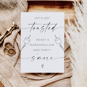 May include: White card with black text that reads "Let's get toasted. Roast a marshmallow and party s'more."  There are two simple drawings of marshmallows on sticks.  The card is on a wooden tray with a beige fabric and a white fluffy plant.