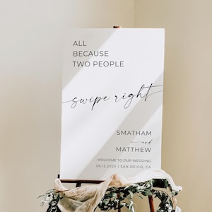 Pode incluir: Uma placa de casamento branca em um cavalete de madeira, decorada com vegetação e tecido. A placa diz "ALL BECAUSE TWO PEOPLE swipe right SMATHAM and MATTHEW WELCOME TO OUR WEDDING 06.12.202X | SAN DEIGO, CA."