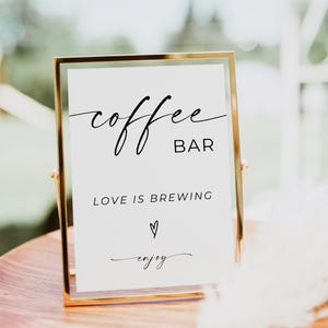 Könnte beinhalten: Ein goldgerahmtes Schild mit der Aufschrift "coffee bar" in Schreibschrift. Darunter steht "Love is brewing" mit einem Herzsymbol und dem Wort "enjoy". Das Schild steht auf einer Holzoberfläche, wahrscheinlich für eine Hochzeit oder Veranstaltung.