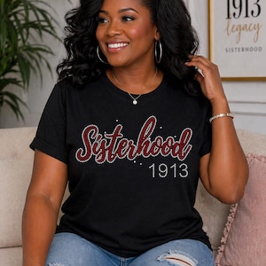 Puede incluir: Camiseta negra con la palabra "Sisterhood" en letras rojas y pedrería, y el número "1913" en blanco. La camiseta se usa con vaqueros azules. Una impresión enmarcada detrás de la persona dice "1913 legacy sisterhood."