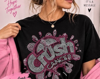 Może przedstawiać: Czarny t-shirt z grafiką wykonaną z różowych i białych cyrkonii, tworzącą napis Crush Cancer. Wzór zawiera motyw rozprysku otaczający tekst. T-shirt jest noszony z różową kurtką.