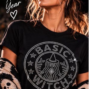 May include: A black short-sleeved t-shirt featuring a circular graphic made of clear rhinestones. The design includes the text "BASIC WITCH" surrounding a central figure wearing a witch hat, accented by small star shapes within the circle.