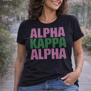 以下が含まれることがあります： ピンクとグリーンのラインストーンで「ALPHA KAPPA ALPHA」の文字が入った黒いTシャツ。Tシャツはブルージーンズと組み合わせています。画像には「Rhinestone Fashion MX」の文字も含まれています。