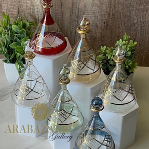 May include: Six decorative glass bottles with gold accents and stoppers. The bottles are arranged on white hexagonal bases. The bottles are clear glass with gold geometric patterns. The bottles are red, beige, clear, blue, and green.