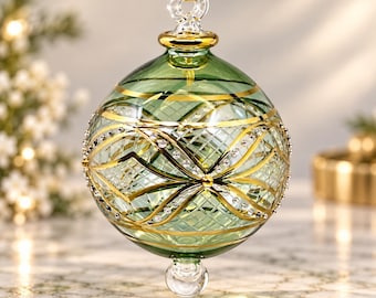 May include: A spherical glass bauble in a translucent sage green hue, featuring intricate gold-toned crisscross lines and delicate silver-coloured bead accents. The bauble is topped with a clear glass loop and a yellow ribbon for hanging.