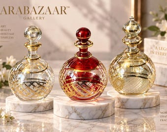 May include: Three round glass perfume bottles with stoppers, featuring gold-tone cross-hatch patterns. The bottles are clear, red, and gold, each resting on a white marble base. Text includes ARABAZAAR GALLERY, ART BEAUTY HERITAGE TIMELESS, A LEGACY IN EVERY SCENT, and MORE THAN A FRAGRANCE.