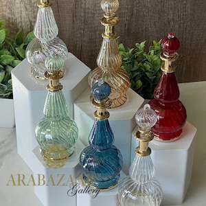May include: Six decorative glass bottles with gold accents and textured surfaces, displayed on white hexagonal platforms. The bottles are in various colours, including clear, green, blue, and red.