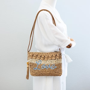 May include: A straw shoulder bag with a brown leather strap and a blue embroidered "Love" on the front. The bag has a tassel with a wooden bead.