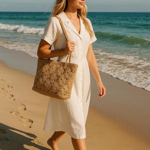 May include: A woman wearing a white button-down dress and carrying a woven tote bag walks on a sandy beach. The tote bag has a floral pattern. The ocean and sky are visible in the background.