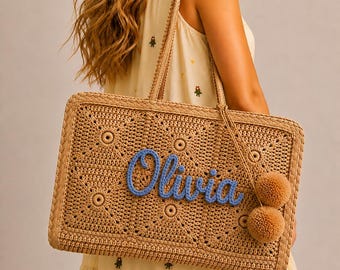 May include: A rectangular straw tote bag featuring a crocheted square pattern in a natural tan color. The front is personalized with the name Olivia in raised blue yarn lettering and includes two matching tan pom-poms attached to the side strap.