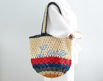 Può includere: Una borsa a mano in crochet con una base marrone chiaro e strisce colorate di rosso, blu e giallo. La borsa ha la parola "Love" ricamata in blu sul davanti.