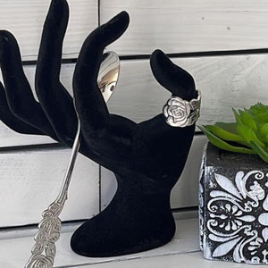 May include: A silver ring with a rose design, displayed on a black velvet hand-shaped ring holder. The ring is on a white background.