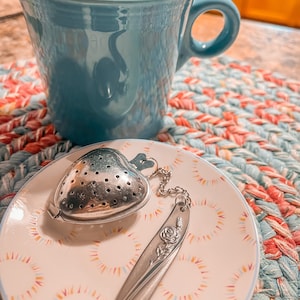 May include: A teal blue ceramic mug with a handle sits on a colourful woven placemat next to a white saucer with a silver heart-shaped tea infuser and a silver tea spoon with a rose design.