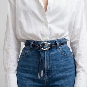 May include: A black leather belt with a silver-toned buckle is worn with blue jeans and a white button-down shirt. The belt has a unique design, with a knot and dangling ends.