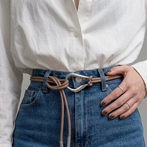May include: A brown leather belt with a silver buckle and a knotted design. The belt is worn with blue denim jeans and a white button-down shirt. The belt has a unique, stylish appearance.