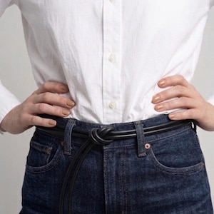 May include: A black leather belt tied in a knot, worn with dark wash denim jeans and a white button-down shirt. The belt has a minimalist design, with silver-toned metal tips.