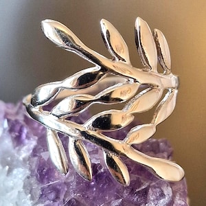 May include: A silver-colored ring with a leaf design. The ring features multiple leaves arranged in a wrap-around style. The ring is displayed on a purple crystal surface, with a blurred background.