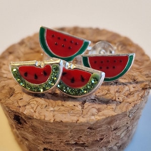 Puede incluir: Pendientes plateados con forma de rodaja de sandía. Cada pendiente presenta una rodaja de sandía de esmalte rojo con detalles de semillas negras y un borde verde. Dos pendientes tienen detalles de cristal verde. Los pendientes se muestran sobre una superficie de corcho.