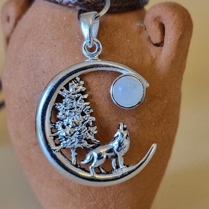 May include: A silver-toned pendant shaped like a crescent moon featuring a textured pine tree and a howling wolf silhouette. A small, round, translucent white stone is set at the top of the crescent. The pendant hangs from a dark brown cord.