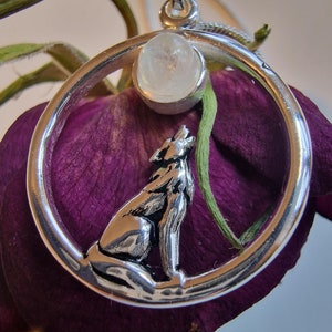 May include: A silver pendant necklace featuring a wolf howling at the moon. The moon is a clear, round, white gemstone set in a silver circle. The pendant is on a purple flower.