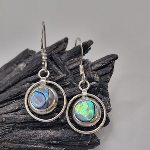 May include: Silver hoop earrings featuring iridescent abalone shell centres. The earrings have a simple design with a hook closure. The abalone shells display colours of blue, green, and purple. A classic jewellery item.