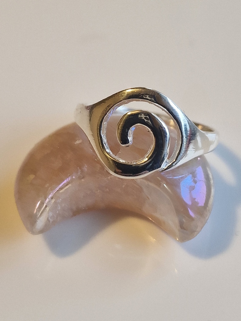 May include: A silver ring with a spiral design. The ring is on a light pink and purple background.