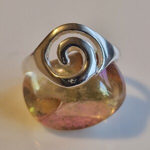 May include: Silver ring with a spiral design. The ring is sitting on a yellow and pink stone.
