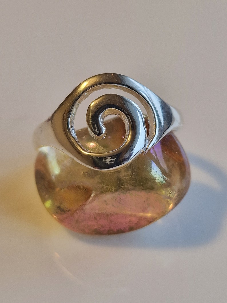May include: Silver ring with a spiral design. The ring is sitting on a yellow and pink stone.