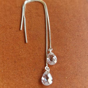 Op de afbeelding: Zilveren oorhangers met twee heldere, druppelvormige kristallen hangers. De oorbellen worden getoond tegen een bruine achtergrond, die hun delicate ontwerp en sprankelende kristallen benadrukt.