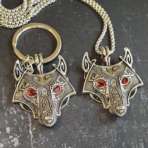 May include: A silver-toned metal keyring and a matching pendant necklace, both featuring a stylised wolf head design with intricate engraved patterns and two vibrant red gemstone eyes set into the metal, connected by a silver box chain.