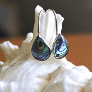 May include: Silver teardrop earrings featuring iridescent abalone shell inlays. The shell's colours include blue, green, and purple. The earrings are presented on a white, textured surface.