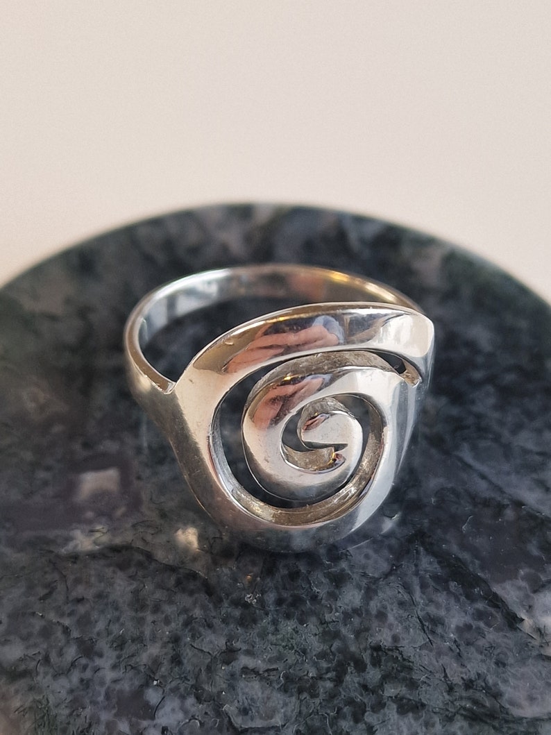 May include: A silver ring featuring a spiral design. The ring is displayed on a dark grey background.