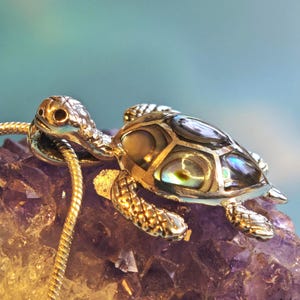 May include: A gold-coloured turtle pendant with iridescent shell details, resting on a purple crystal. The pendant is attached to a thin, gold-coloured chain. The background is a soft gradient of blue and green.