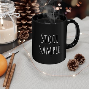 Può includere: Tazza da caffè nera con testo bianco che recita "STOOL SAMPLE". La tazza è appoggiata su una superficie grigio chiaro con una ghirlanda di luci e pigne.