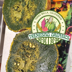 May include: Two halves of a green-skinned squash, cut to reveal the interior. The squash is on a floral cutting board with an orange knife. A circular logo reads "Terra Bella, California" and "Tremesino Organics."