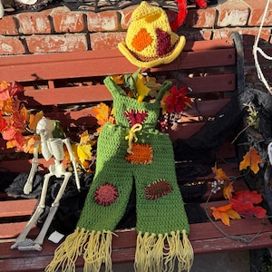 May include: A green crochet overall with yellow fringe at the ankles, featuring orange, brown, and maroon patches, and a matching yellow crochet hat with orange and maroon accents. The overall has a yellow drawstring at the waist.